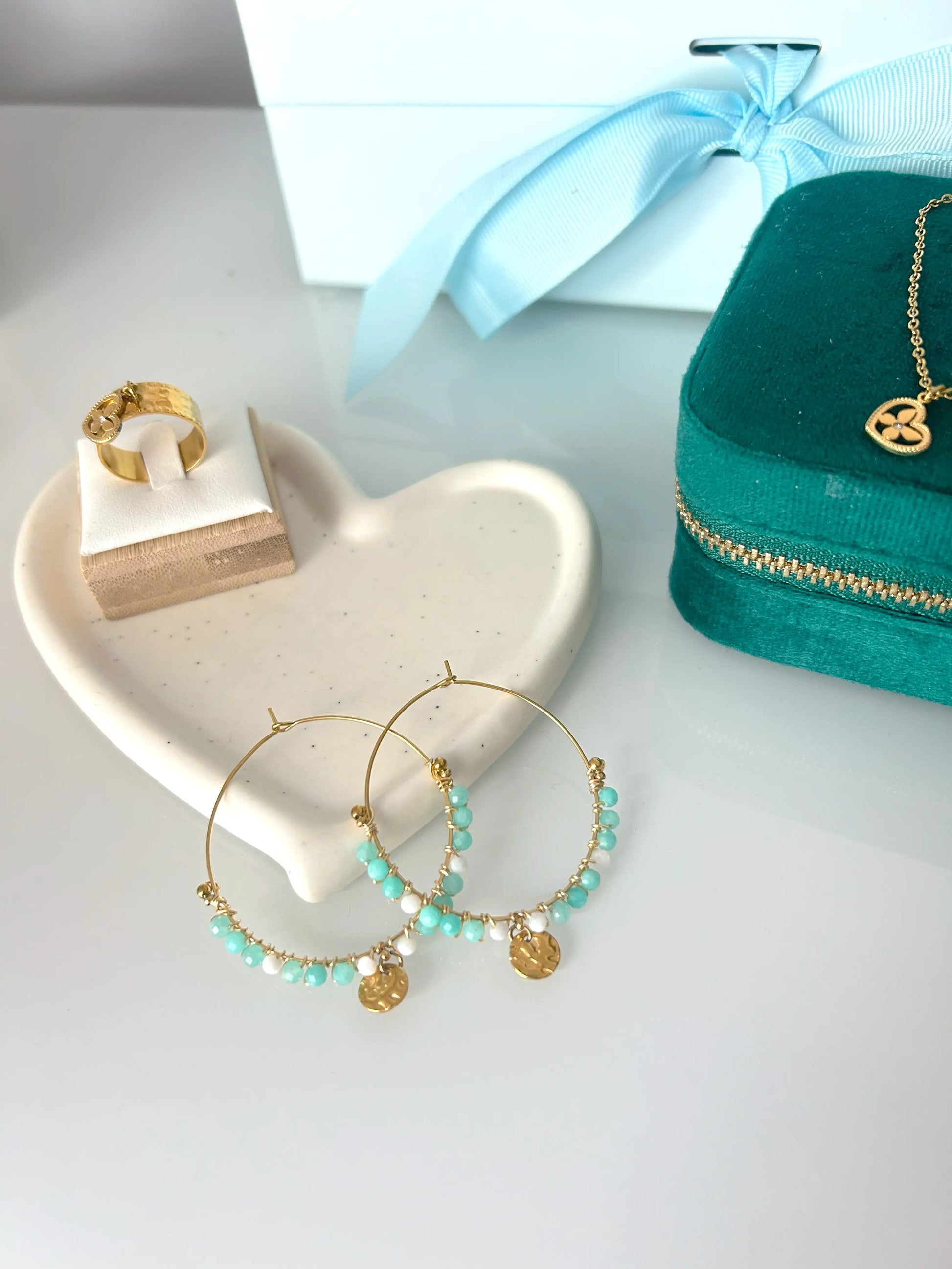 Boucles d'oreilles créoles en acier inoxydable doré posée sur une coupelle coeur en céramique blanche, posée sur une table décorée d'une boîte à bijoux vert émeraude avec une bague et un collier doré avec un pendentif coeur papillon. Bijou orné de perles de verre blanc et turquoise avec un sequin rond martelé suspendu. 