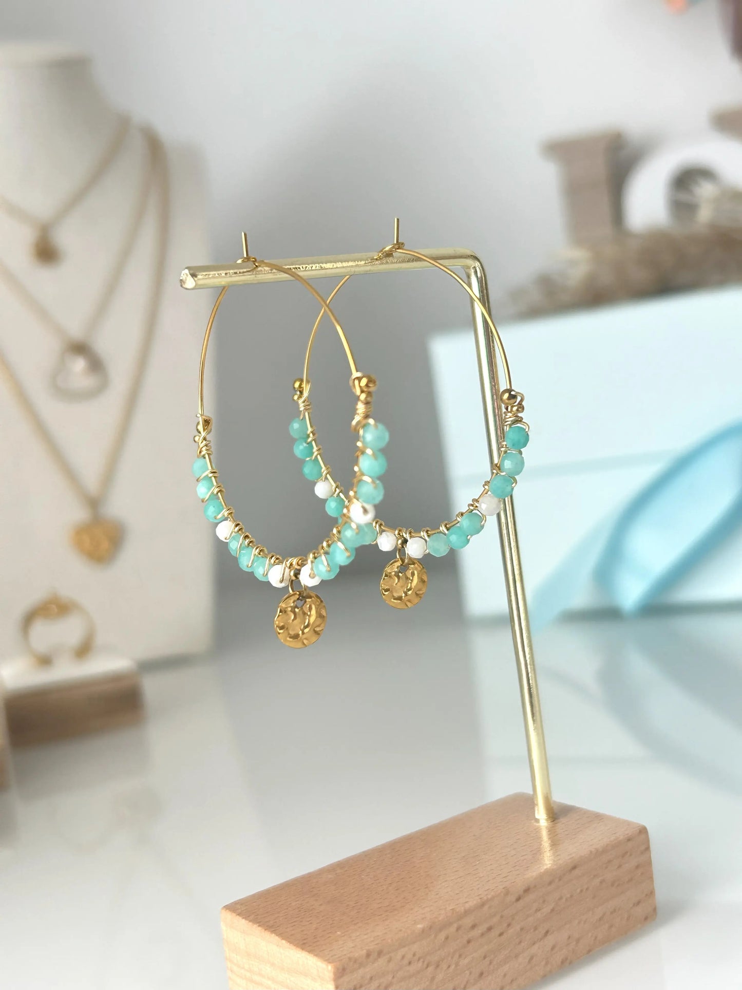 Boucles d'oreilles créoles en acier inoxydable doré suspendues sur un support métallique doré et en bambou posée sur une table en verre trempée. Bijou orné de perles de verre blanc et turquoise avec un sequin rond martelé.