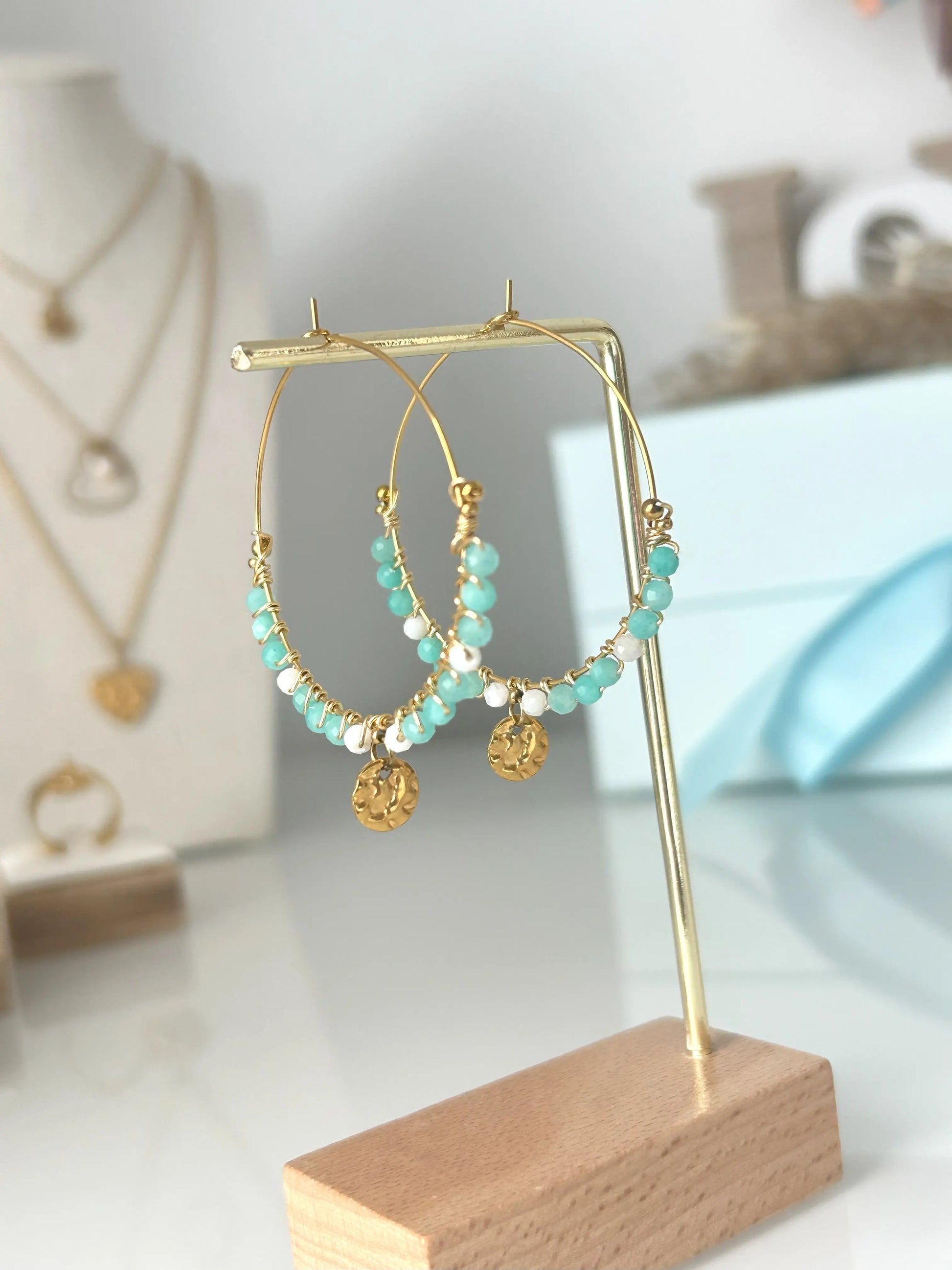 Boucles d'oreilles créoles en acier inoxydable doré suspendues sur un support métallique doré et en bambou posée sur une table en verre trempée. Bijou orné de perles de verre blanc et turquoise avec un sequin rond martelé.