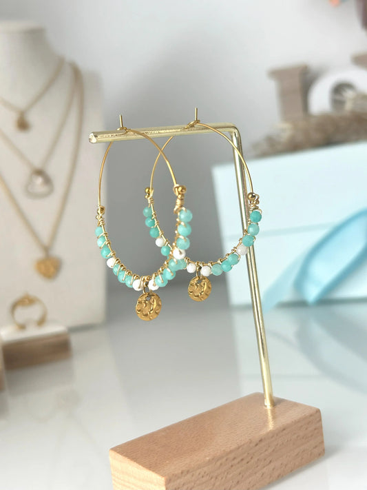 Boucles d'oreilles créoles en acier inoxydable doré suspendues sur un support métallique doré et en bambou posée sur une table en verre trempée. Bijou orné de perles de verre blanc et turquoise avec un sequin rond martelé.