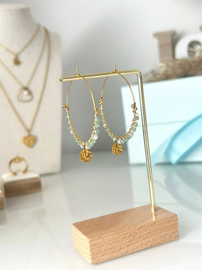 Boucles d'oreilles créoles et sequin martelé suspendu en acier inoxydable doré. Bijou orné de perles de verre gris et vert. Bijou suspendu à un support métallique doré et en bambou.
