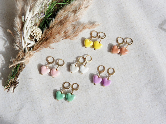Boucles d'oreilles créoles en acier inoxydable doré, décorées d'une perle cœur en céramique colorée jaune, marron, mauve, blanc, vert et rose. Bijoux posés à plat sur une nappe beige en coton et décorée d'un mini-bouquet de fleurs.