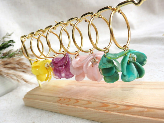 Boucles d'oreilles créoles en acier inoxydable doré suspendues à un support métallique doré et en bambou posé sur une nappe beige. Boucles décorées de pétales colorées jaune, violet, rose et vert à effet marbré, formant un bouquet floral suspendu à la créole. 