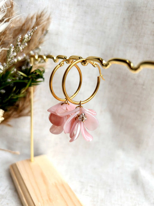 Boucles d'oreilles créoles en acier inoxydable doré suspendues à un support métallique doré et en bambou posé sur une nappe beige. Boucles décorées de pétales colorées rose à effet marbré, formant un bouquet floral suspendu à la créole. 
