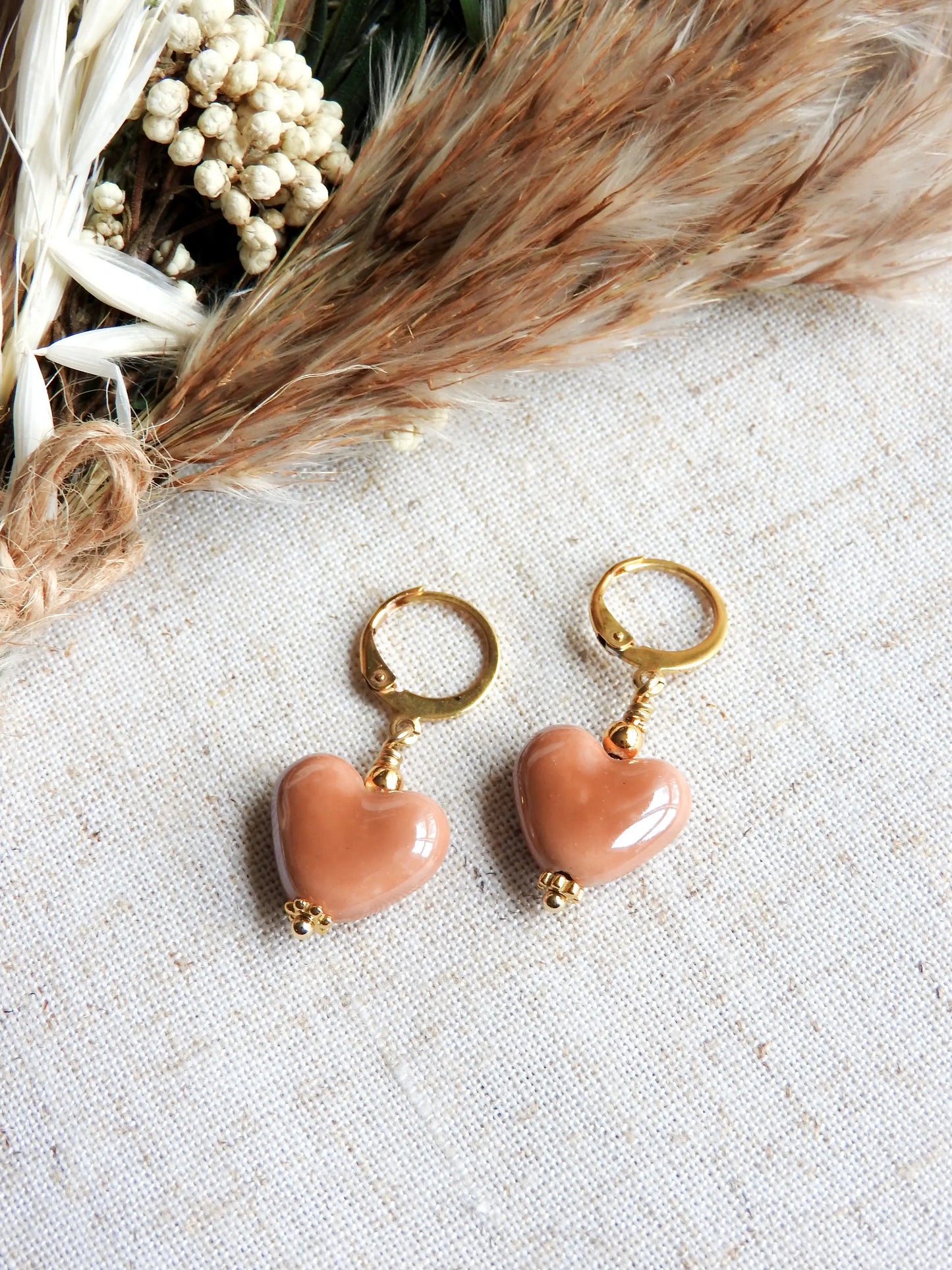 Boucles d'oreilles dormeuses créoles en acier inoxydable doré, décorées d'une perle cœur en céramique marron. Bijou posé à plat sur une nappe beige en coton et décorée d'un mini-bouquet de fleurs.