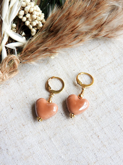 Boucles d'oreilles dormeuses créoles en acier inoxydable doré, décorées d'une perle cœur en céramique marron. Bijou posé à plat sur une nappe beige en coton et décorée d'un mini-bouquet de fleurs.