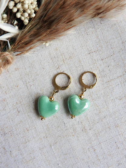 Boucles d'oreilles dormeuses créoles en acier inoxydable doré, décorées d'une perle cœur en céramique verte. Bijou posé à plat sur une nappe beige en coton et décorée d'un mini-bouquet de fleurs.