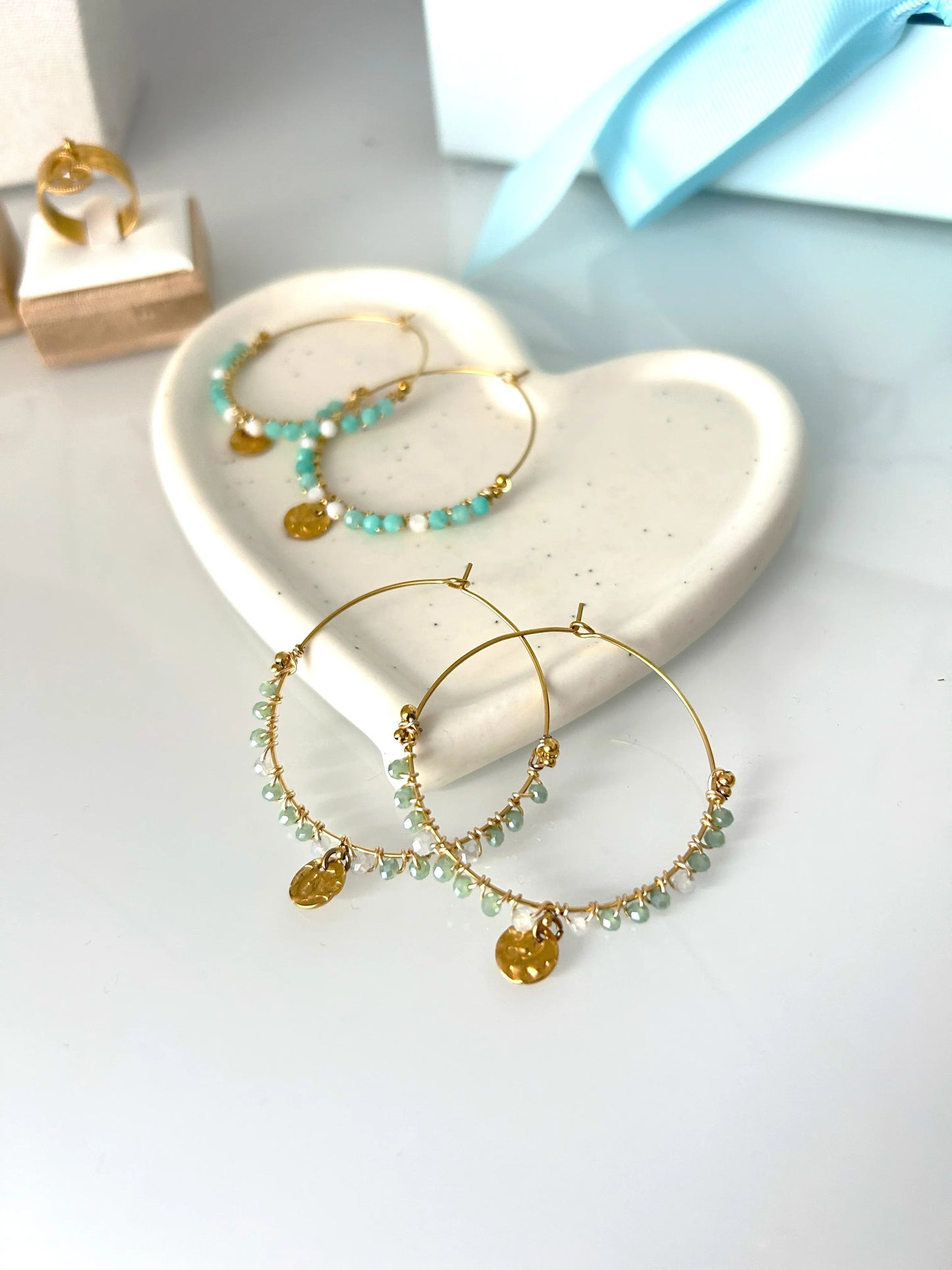 deux paires de boucles d'oreilles créoles en acier inoxydable doré posées à plat sur une coupelle en céramique blanche. Bijoux ornés de perles de verre blanc et turquoise pour une paire, et vert et gris pour l'autre paire avec un sequin rond martelé en suspension.