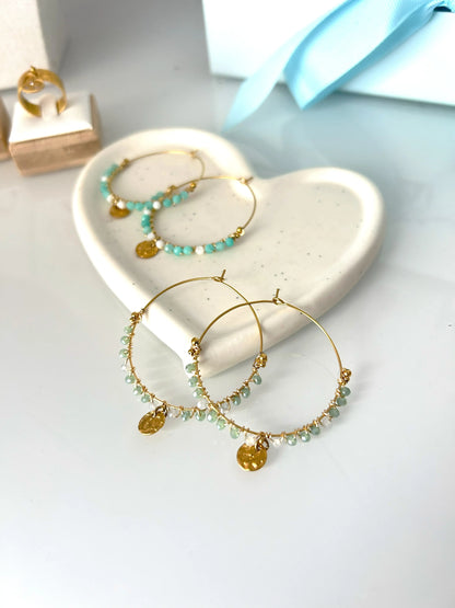 deux paires de boucles d'oreilles créoles en acier inoxydable doré posées à plat sur une coupelle en céramique blanche. Bijoux ornés de perles de verre blanc et turquoise pour une paire, et vert et gris pour l'autre paire avec un sequin rond martelé en suspension.