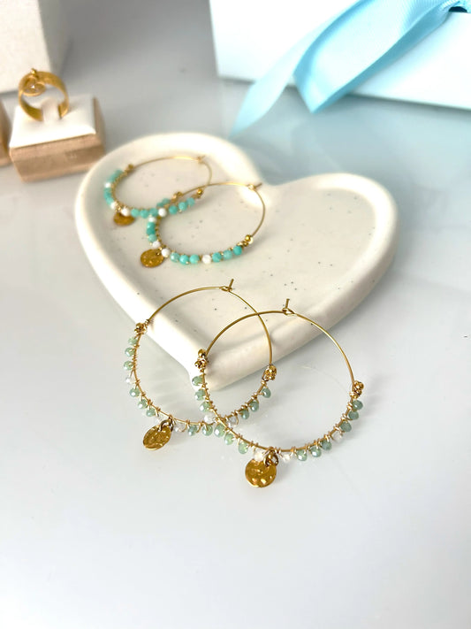 deux paires de boucles d'oreilles créoles en acier inoxydable doré posées à plat sur une coupelle en céramique blanche. Bijoux ornés de perles de verre blanc et turquoise pour une paire, et vert et gris pour l'autre paire avec un sequin rond martelé en suspension.