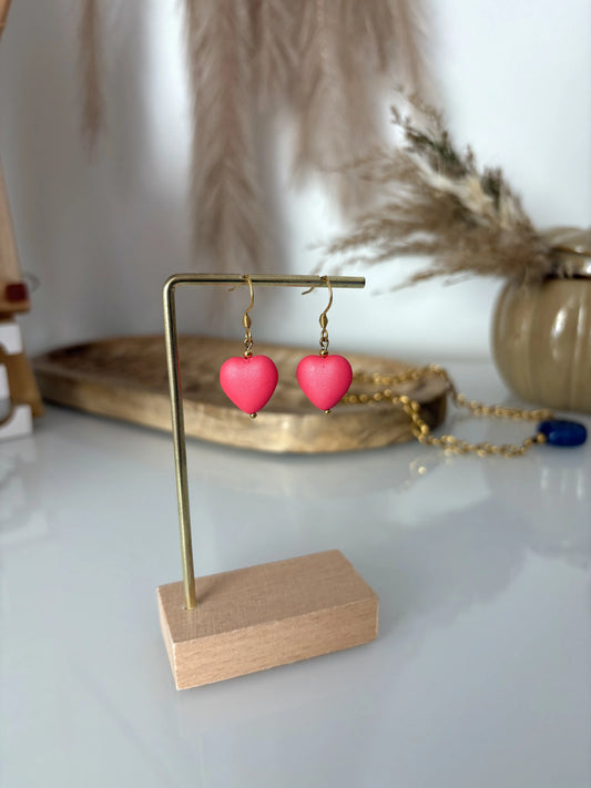 Boucles d'oreilles crochet en acier inoxydable doré avec une perle coeur couleur hibiscus en suspension. Bijou posé sur un support métallique doré et bambou. 
