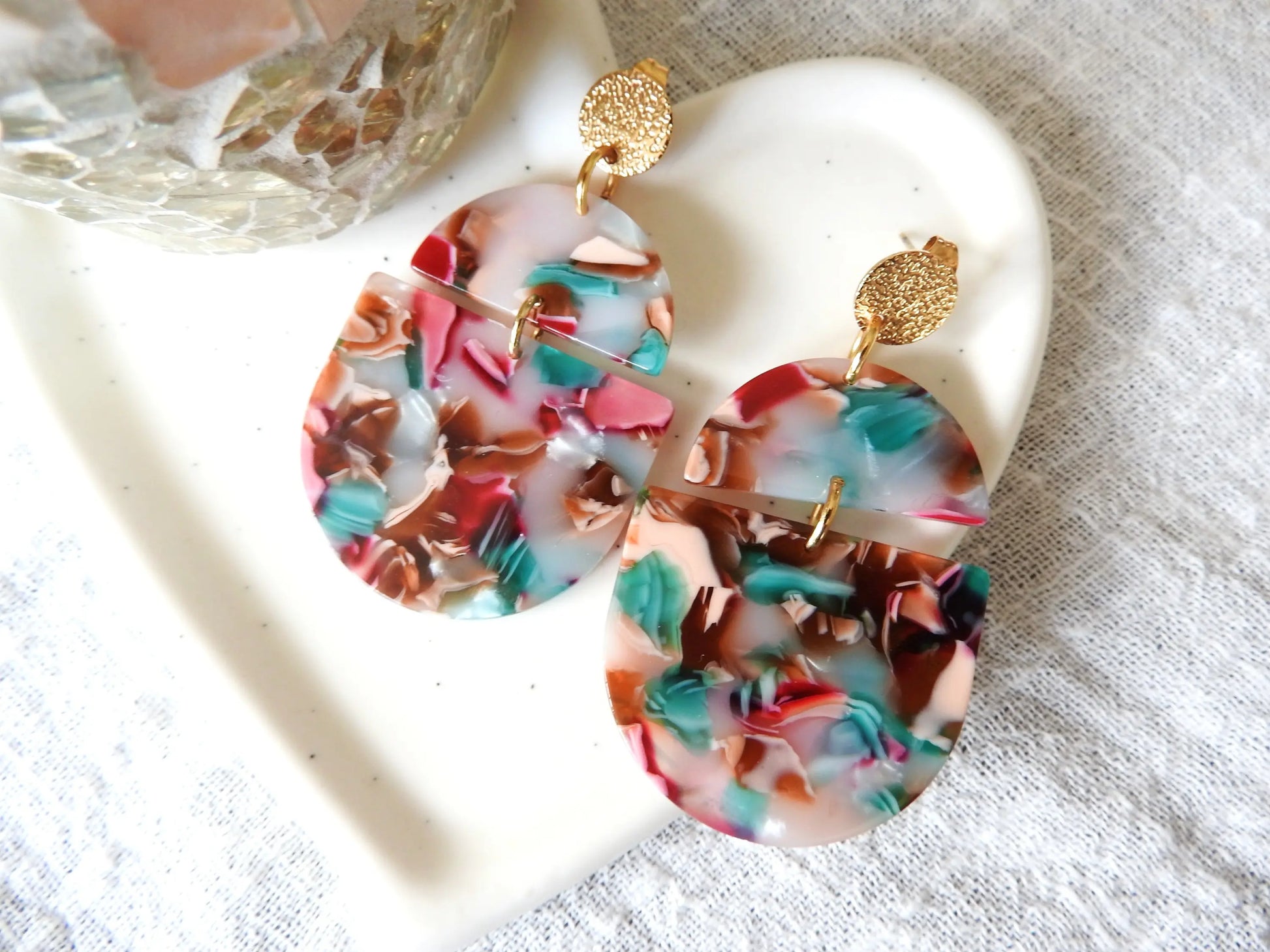 boucles d'oreilles clous disque martelé en acier inoxydable doré. Bijou orné de deux formes géométriques, demi-cercle et demi-ovale multicouleurs. Bijou posé sur une coupelle coeur en céramique blanche.