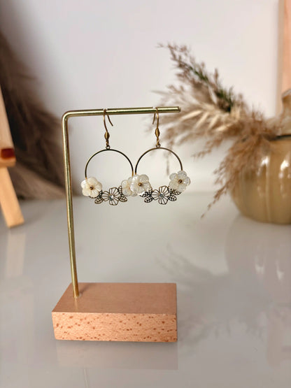 Boucles d'oreilles crochets en acier inoxydable doré, élégantes pour cérémonie. Bijoux ornées de strass et petites fleurs en nacre blanche. Bijoux présentés sur un support métallique doré et bambou posé sur une table en verre.