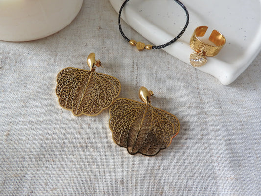 boucles d'oreilles clous en forme de goutte ornées d'un pendentif papillon filigrane en acier inoxydable doré. Bijoux posés sur une nappe beige en coton avec d'autres bijoux posés sur une coupelle en céramique : bracelet cordon noir à fil argenté avec des perles doré dont une perle coeur gravée de la lettre M et une bague anneau réglable à effet martelé avec une breloque coeur ornée de pierres en oxydes de zirconium.