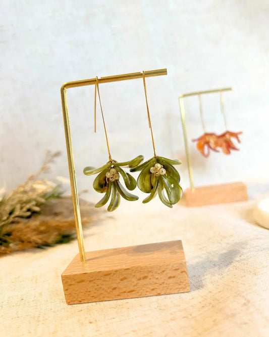 Boucles d'oreilles hameçon en acier inoxydable doré ornées de pétales vert olive formant un bouquet floral. Bijoux agrémentés de perles de verre et acier doré, boucles suspendues sur un présentoir métallique doré et bambou posé sur une nappe beige décorée d'une brindille.