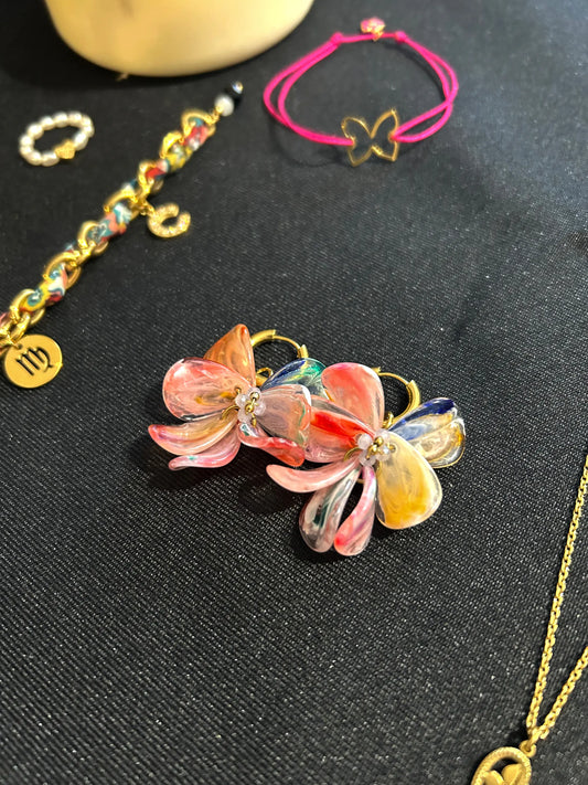 boucles d'oreilles mini-créoles en acier inoxydable doré posées à plat sur une nappe noire. Boucles d'oreilles composées de pétales en acrylique multicouleurs rose, fuchsia, jaune, orange, bleu et vert formatnt un bouquet floral. A côté, bracelet cordon en nylon tressé fuchsia orné d'un papillon en acier doré.