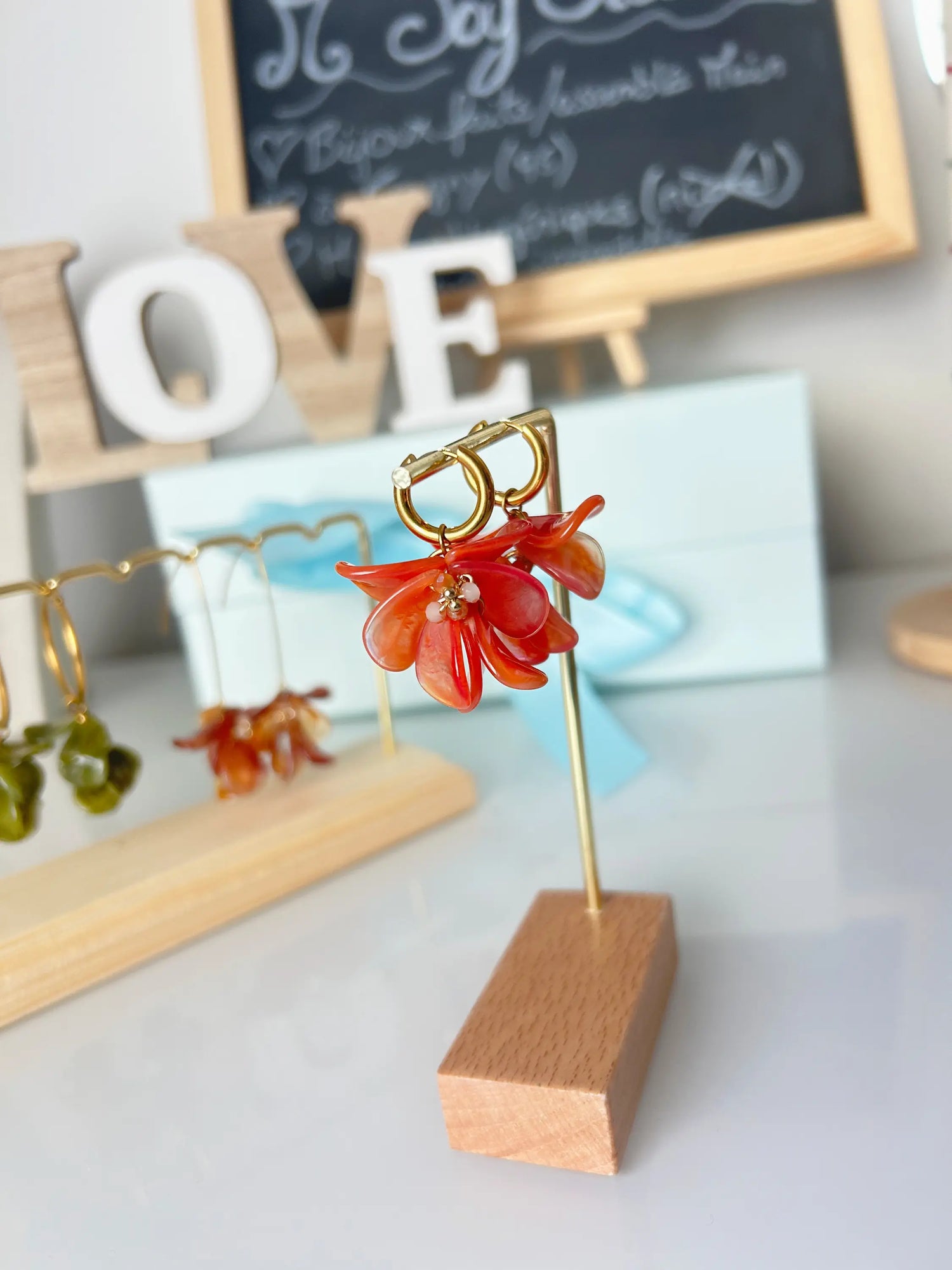 boucles d'oreilles mini-créoles en acier inoxydable doré suspendues à un présentoir métallique doré et bambou. Bijou orné de pétales formant un bouque floral en acrylique de couleur orange marbré. 