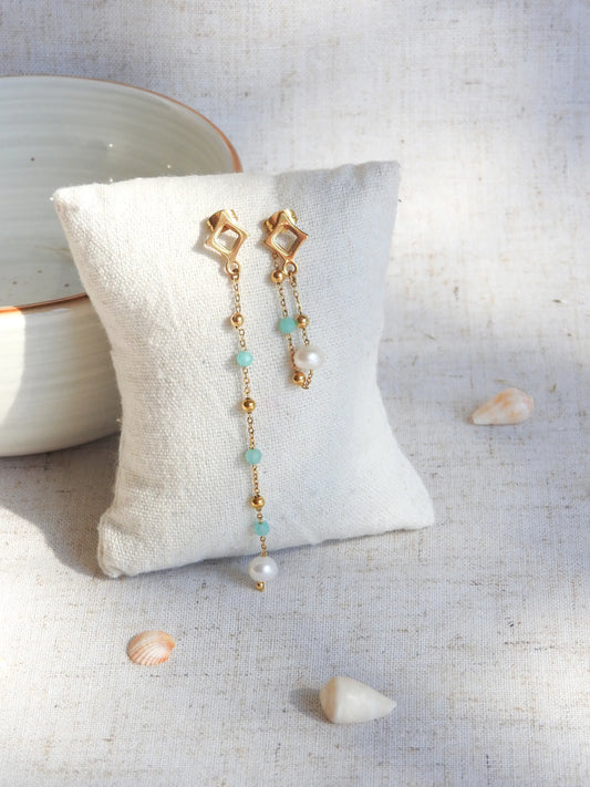 boucles d'oreilles clous losange en acier inoxydable doré. Chainettes pendantes asymétriques perlées en acier doré et perles de verre turquoise et perles d'eau douce blanche. Bijoux piqués sur un mini-coussin beige, contre un bol nu céramique, sur une table décorée de coquillages.