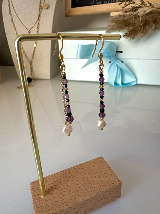 Boucles d'Oreilles Duo Pendantes en perles de verre violettes et noires et Perle d'eau douce suspendue à une chaînette. Bijou présenté sur un support métallique doré et bambou.