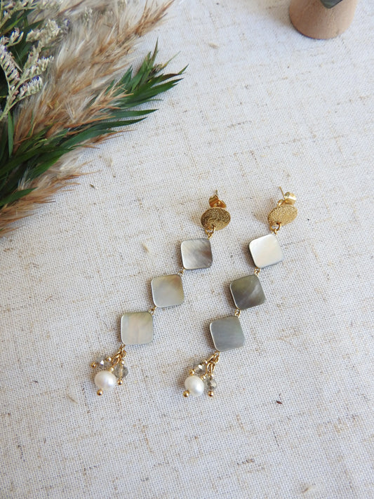 boucles d'oreilles clous à disque martelé ornées de perles carré en nacre en suspension et finissant avec des perles d'eau douce blanche et en verre grises. Bijoux posés à plat sur une nappe beige.