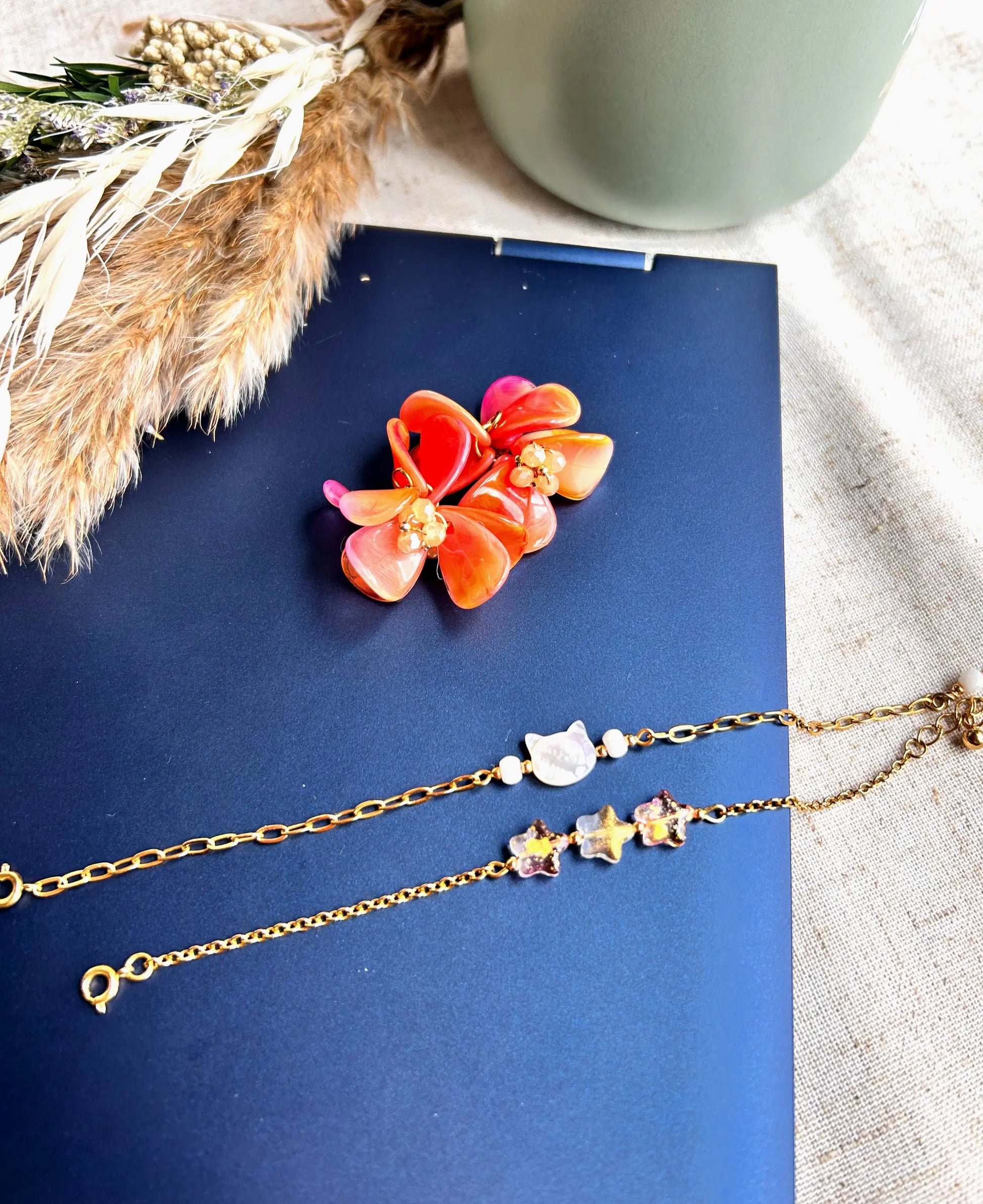 Suspensions pour créoles en acier inoxydable assemblées à la main. Bouquet floral réalisé à partir de pétales en acrylique orange marbré et de perles de verre colorées et en acier inoxydable doré. Bijoux posés sur le dessus d'un ordinateur avec deux bracelet chaîne en acier inoxydable doré avec une perle chat en nacre blanche et des perles corail en forme d'étoile scintillantes.