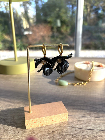 Boucles d'oreilles petites créoles en acier inoxydable doré et ornées de pétales noires superposées et perles de verre. Bijoux présentés sur une barre métallique dorée et en bambou.