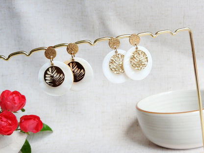 deux paires de boucles d'oreilles clous à disque à effet diamanté décorées d'un double sequin rond, l'un en nacre blanche et l'autre avec une feuille évidée en acier dorée à texture lisse pour la première paire et à texture martelée pour la deuxième paire. 