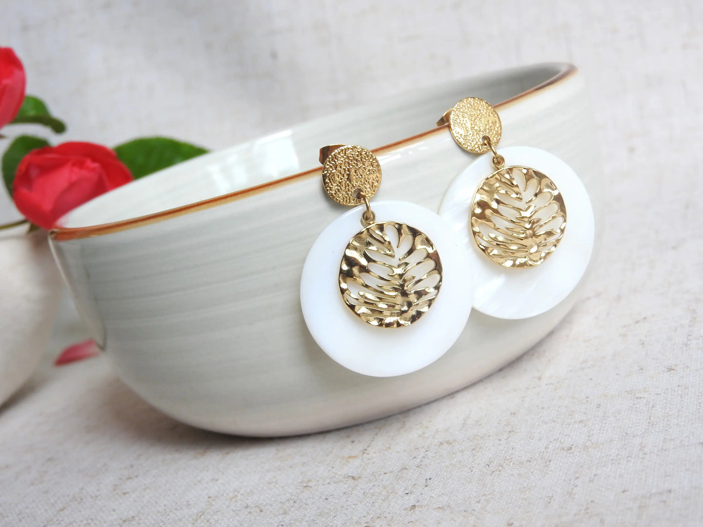 boucles d'oreilles clous à disque effet diamanté en acier doré, ornées d'un double sequin, l'un en nacre blanche et l'autre en médaille rond avec une feuille évidée. Bijou suspendu à un bol en céramique posé sur une nappe beige décorée de roses roses. 