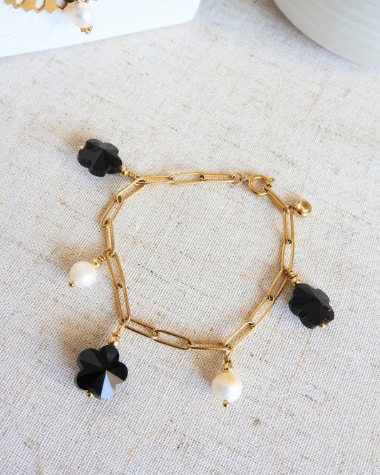 Bracelet posé à plat et fermé sur une nappe beige, bracelet chaine à maille rectangle en acier inoxydable doré, orné de breloques en perles d'eau douce blanche et perles fleurs en verre noire.