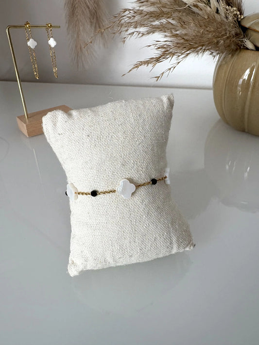 Bracelet chaine en acier inoxydable doré perlés de perles de verre noire ainsi que des perles trèfle en nacre blanche. Bijou présenté sur un mini-coussin beige en coton posé sur une table décorée de brindilles, un pot en céramique et des boucles d'oreilles pendantes.