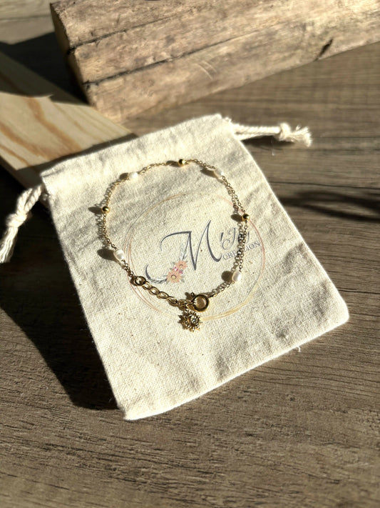 Bracelet à chaîne fine perlé dorée et à perles d'eau douce blanche. Bijou orné d'une pampille soleil à strass et disposé sur un pochon à coton beige siglé de la marque de bijoux M'Joy Creation.