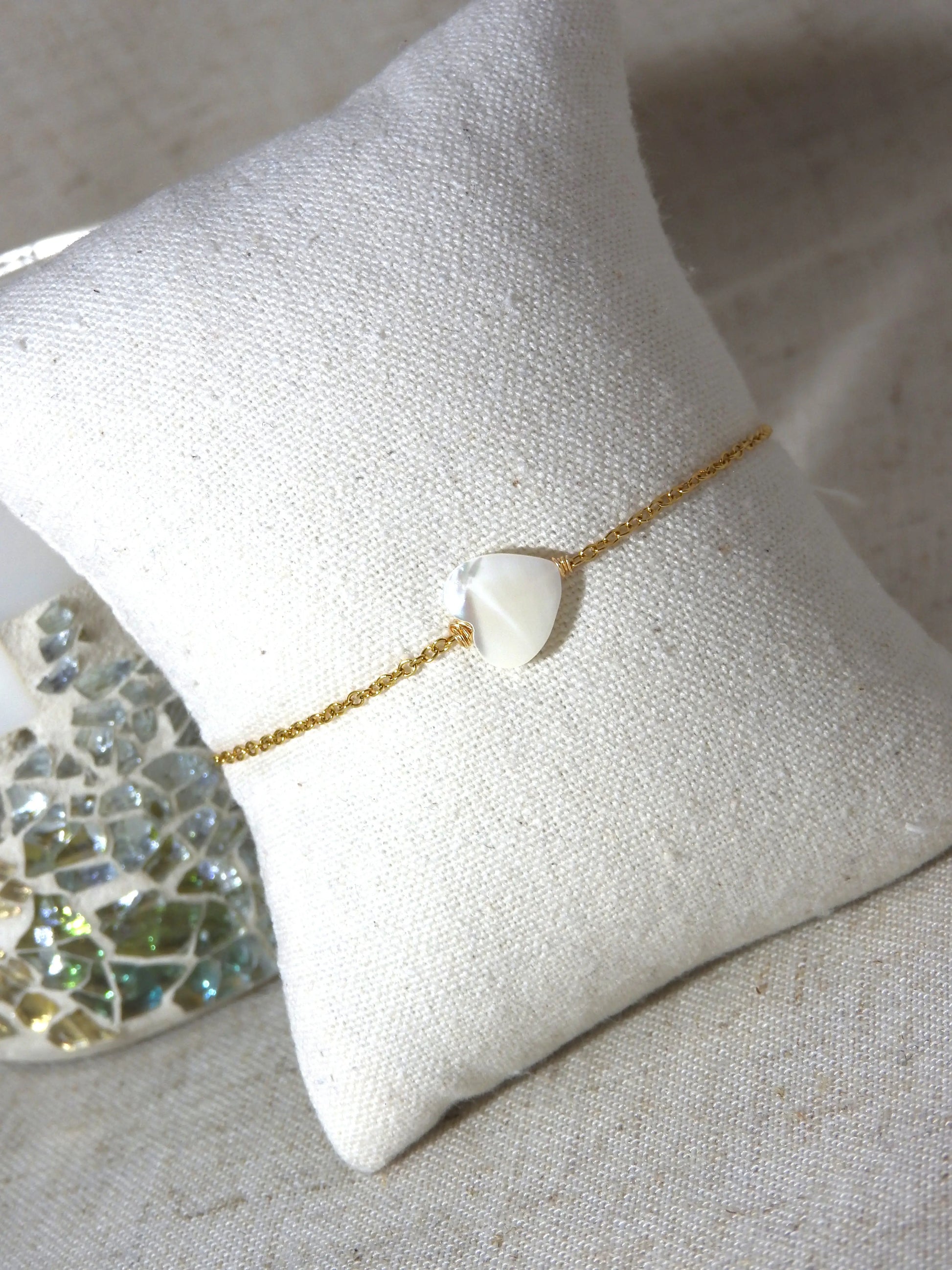 Bracelet chaîne fine en acier inoxydable doré posé sur un mini-coussin beige en coton. Bijou orné d'une perle coeur en nacre blanche.