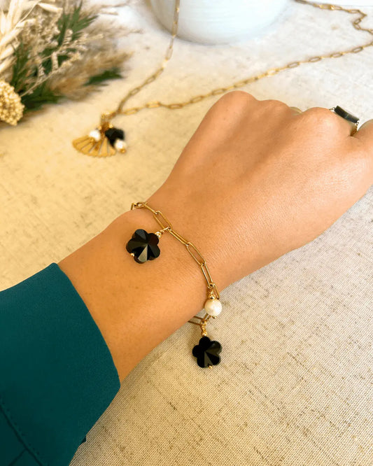Bracelet posé à plat sur une nappe beige, composé d'un collier chaîne mailles rectangle en fond  En avant plan, un bracelet chaine maille rectangle en acier inoxydable doré, orné de breloques en perles d'eau douce blanche et perles fleurs en verre noire. Bijou porté au poignet posé sur la table.
