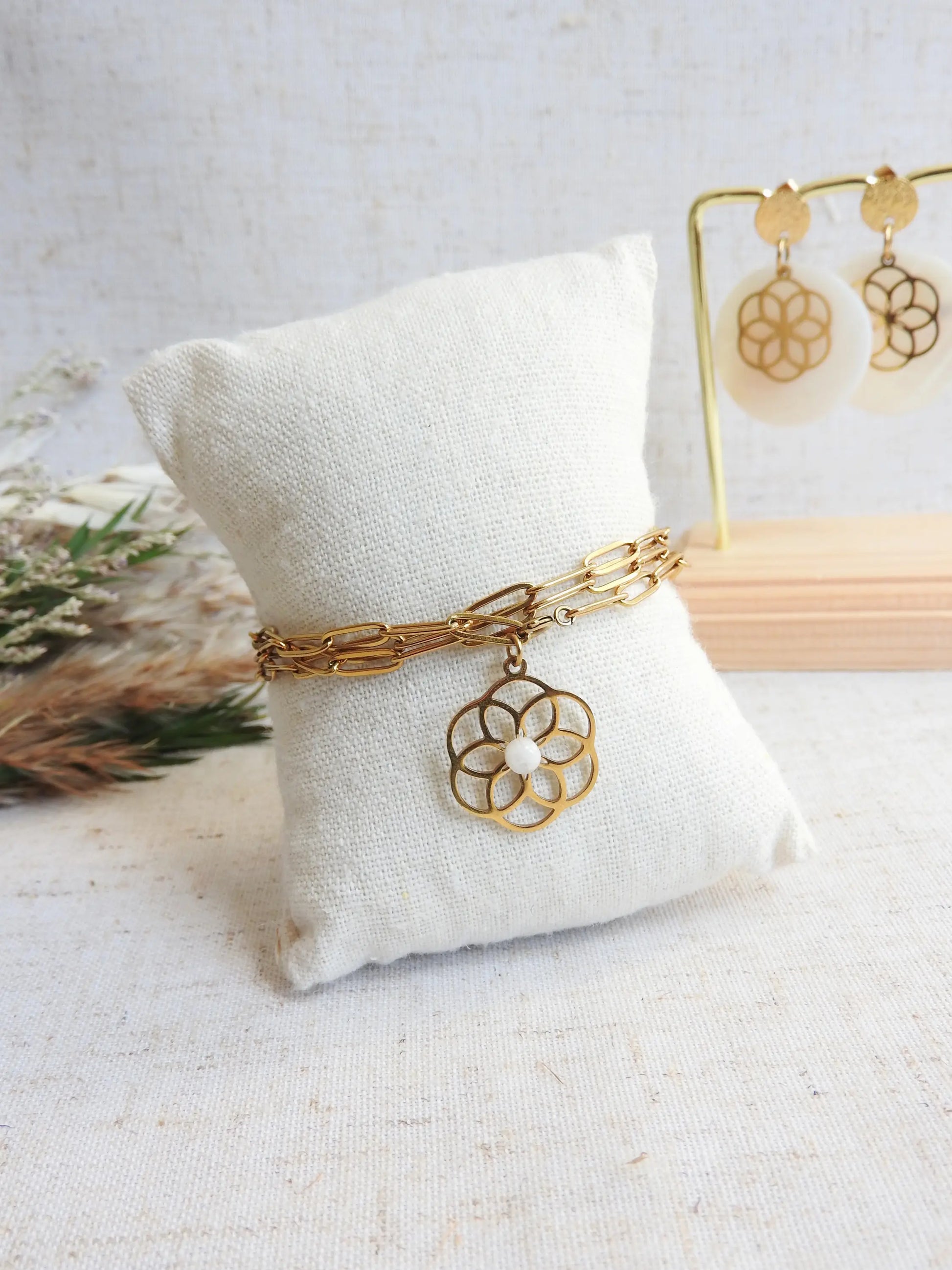 Bracelet chaîne trois tours à maille rectangle en acier inoxydable doré présenté sur un mini-coussin beige. Bijou orné d'un pendentif fleur filigrane en acier doré, orné d'une pierre de lune. 
