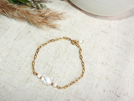 Bracelet chaîne à mailles rectangle orné de perles blanches et une perle chat en nacre blanche. Bijou posé sur une nappe beige décorée d'une brindille.