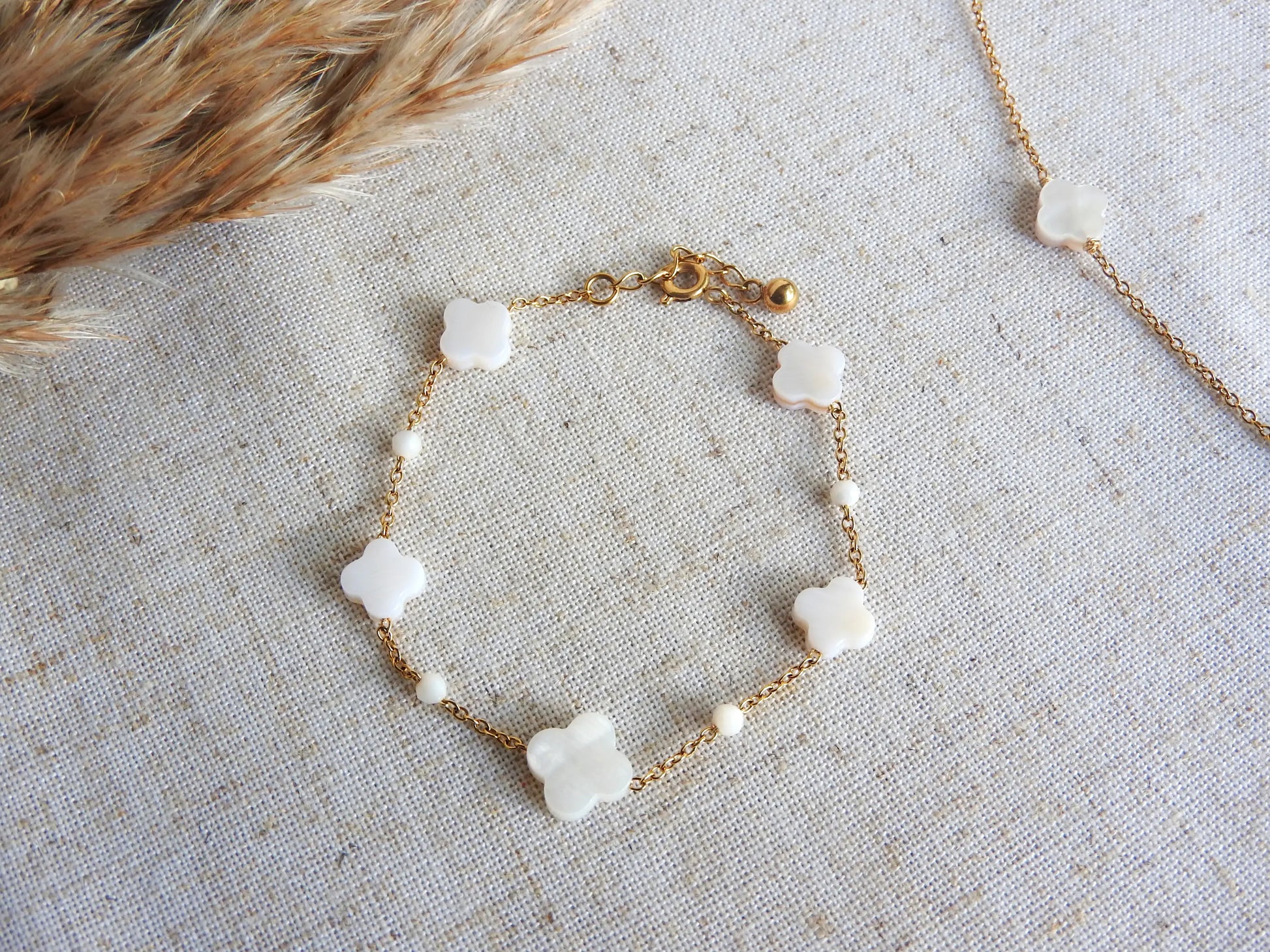 bracelet à chaine perlée en perles de verre blanche et perles trèfle en nacre blanche. Bijou en acier inoxydable doré posé à plat sur une nappe beige décorée d'une brindille.