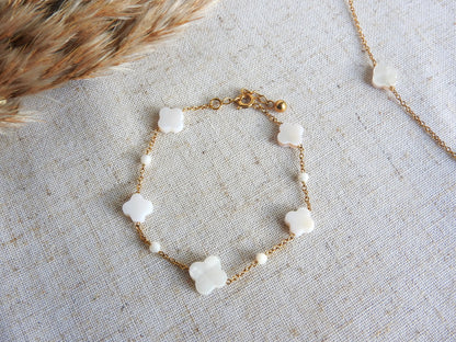 bracelet à chaine perlée en perles de verre blanche et perles trèfle en nacre blanche. Bijou en acier inoxydable doré posé à plat sur une nappe beige décorée d'une brindille.