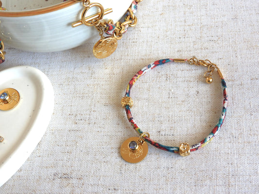 Bracelet cordon liberty Thorpe hill aux multicouleurs florales. Pendentif Fleur et pierre de naissance en acier inoxydable doré. Bijou présenté à plat sur une nappe beige.