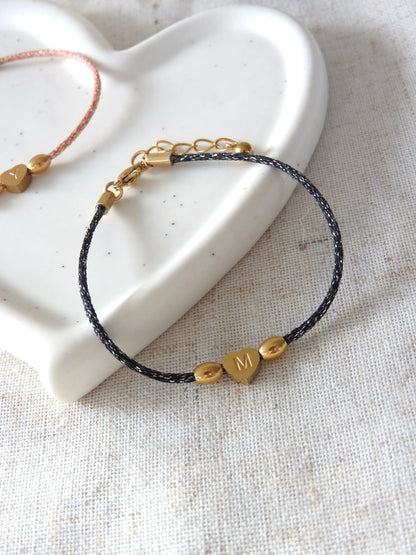 Bracelet cordon noir à fil argenté orné de 3 perles dont une en forme de coeur plat  gravée de la lettre M en acier inoxydable doré. Bijou posé sur une coupelle blanche en céramique.