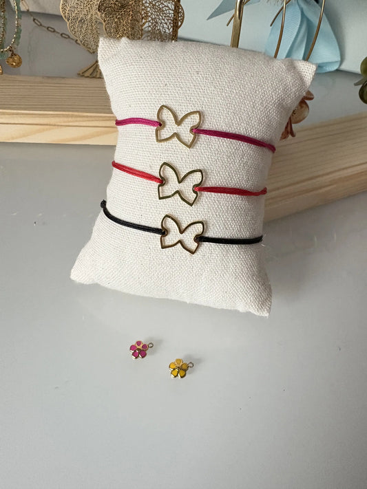 Trois bracelets en nylon tressé de couleurs fuchsia, rouge et noir disposés sur un mini-coussin en coton beige. Bijoux ornés d'un papillon en acier inoxydable doré et de mini-breloques fleur en résine époxy fuchsia et jaune.