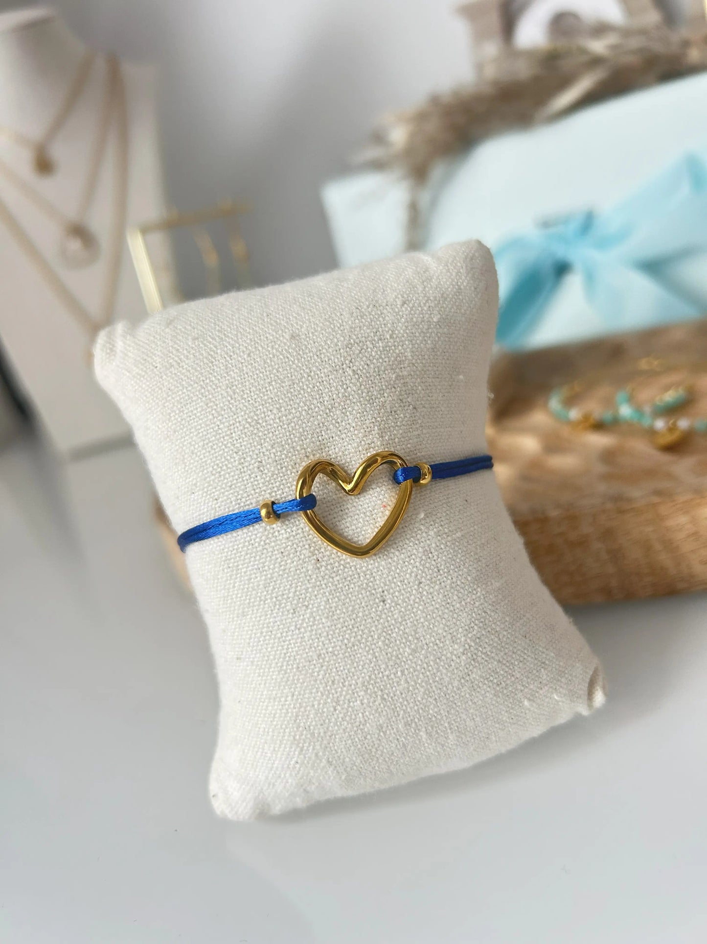 bracelet cordon ajustable en nylon bleu roi orné d'un cœur courbé en acier inoxydable doré. Bijou présenté sur un mini-coussin beige en coton, posé sur une table décorée d'un plateau en bambou.