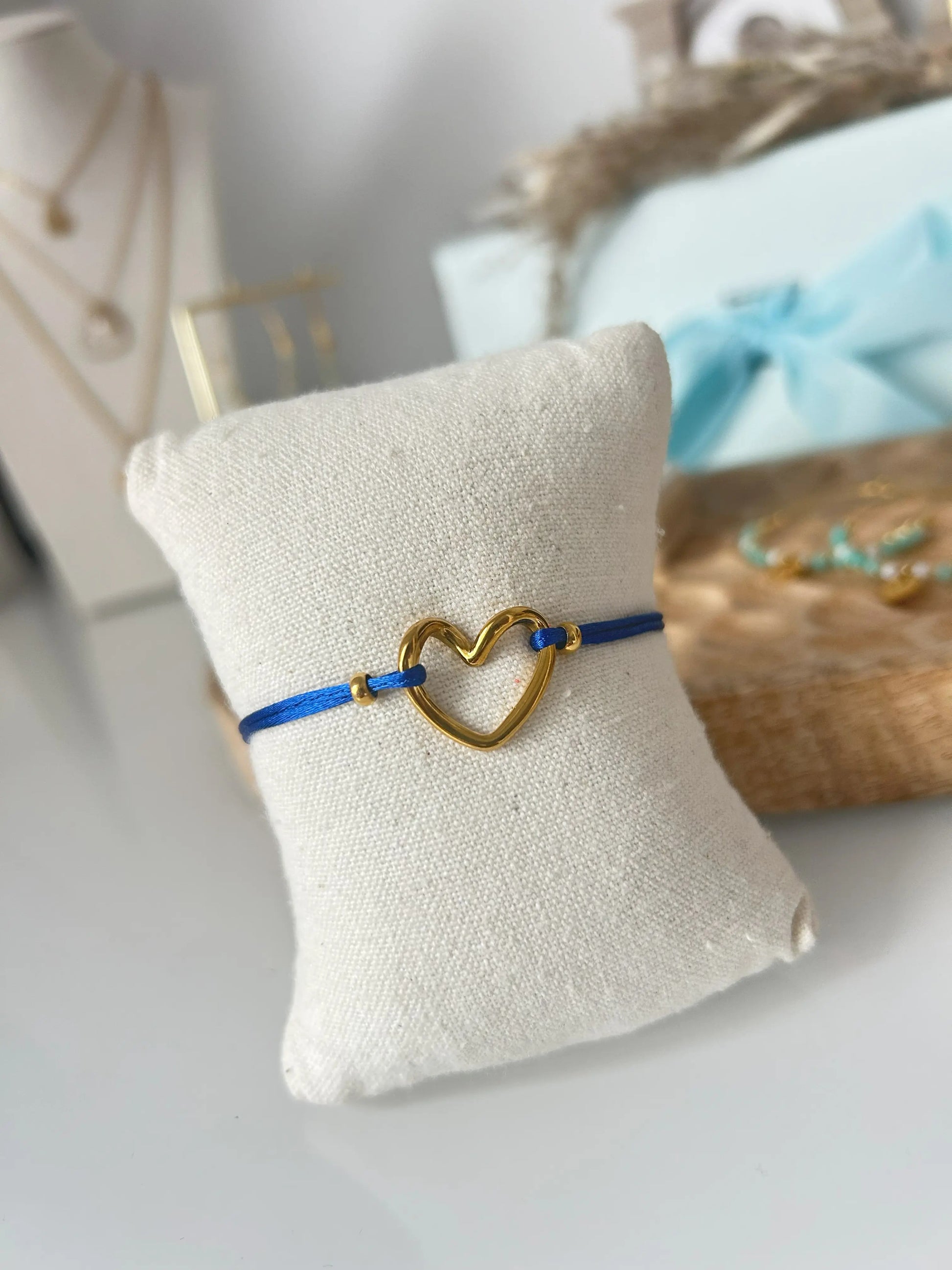 bracelet cordon ajustable en nylon bleu roi orné d'un cœur courbé en acier inoxydable doré. Bijou présenté sur un mini-coussin beige en coton, posé sur une table décorée d'un plateau en bambou.
