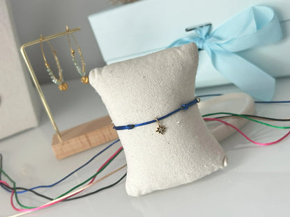 bracelets cordon ajustable en nylon bleu roi ornées d'une pampille étoile du nord avec une pierre en oxyde de zirconium et en acier inoxydable doré. Bijou présenté sur un mini-coussin beige en coton, posé sur une table décorée de cordon en nylon aux couleurs noir, vert électrique, champagne, rose fuchsia et bleu roi.