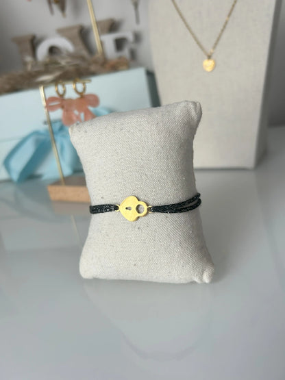 bracelet cordon scintillant noir à fil argenté présenté sur un mini-coussin beige en coton.  Bijou orné d'un cadenas coeur en acier inoxydable doré. En fond un buste avec un collier doré et une paire de boucles d'oreilles dorées et saumon sur un présentoir en bambou. 
