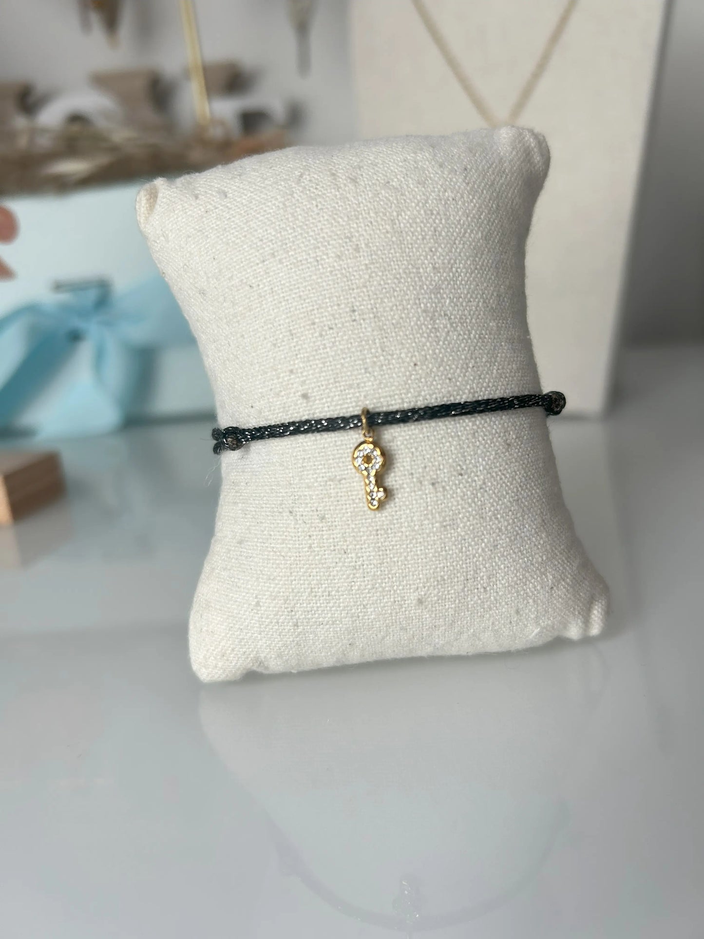 bracelet cordon scintillant noir à fil argenté présenté sur un mini-coussin beige en coton.  Bijou orné d'une clé avec strass en acier inoxydable doré. En fond un buste avec un collier doré et une boite bleu ciel.