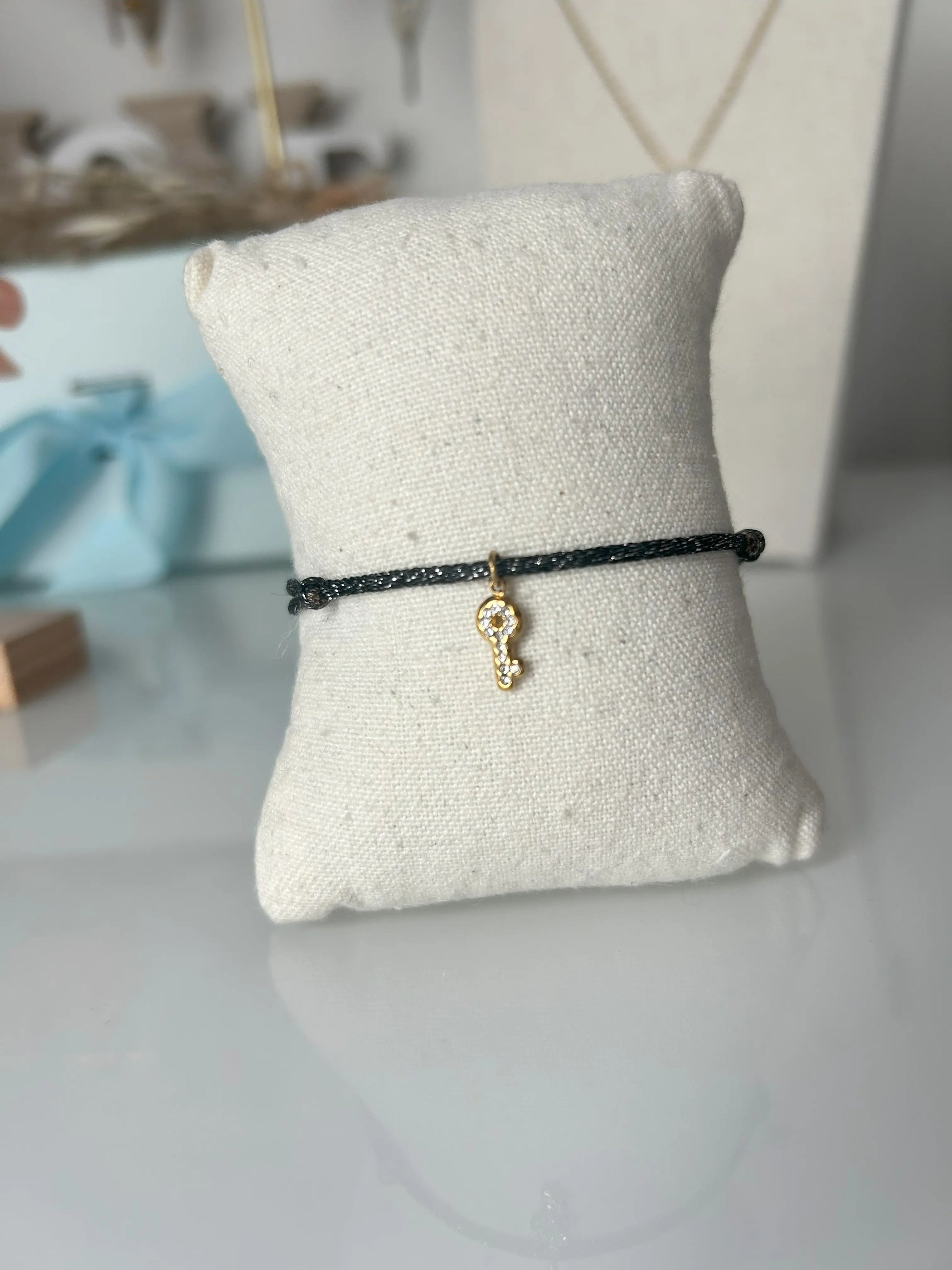 bracelet cordon scintillant noir à fil argenté présenté sur un mini-coussin beige en coton.  Bijou orné d'une clé avec strass en acier inoxydable doré. En fond un buste avec un collier doré et une boite bleu ciel.