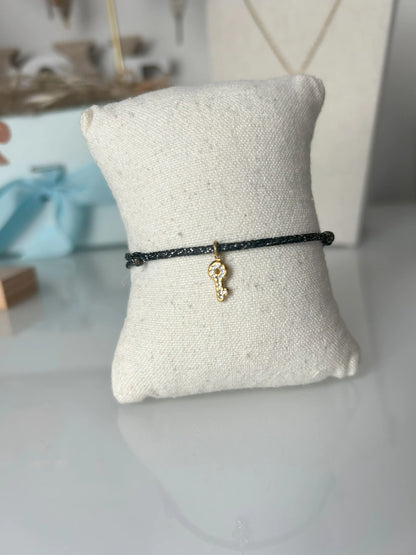 bracelet cordon scintillant noir à fil argenté présenté sur un mini-coussin beige en coton.  Bijou orné d'une clé avec strass en acier inoxydable doré. En fond un buste avec un collier doré et une boite bleu ciel.