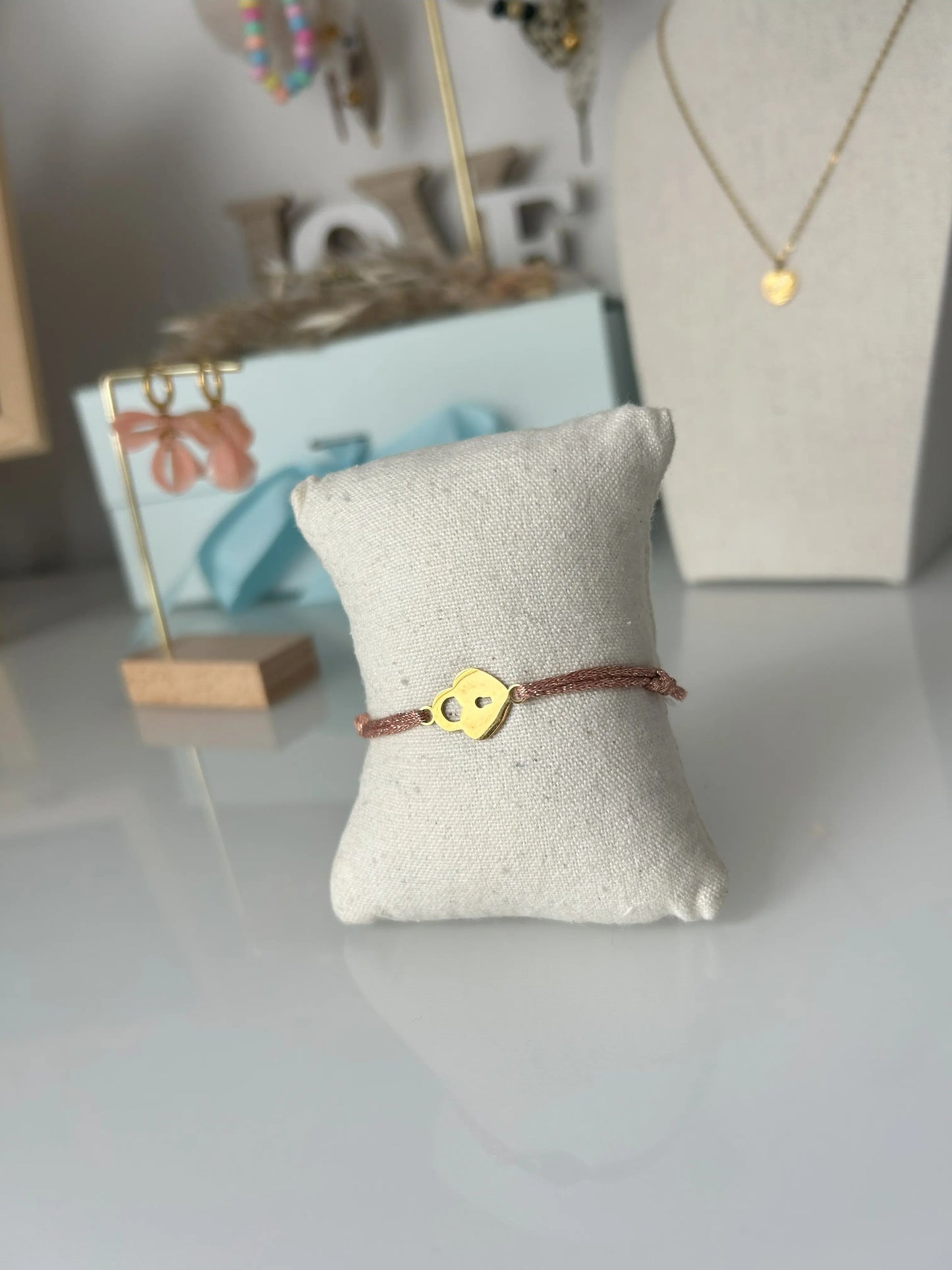 bracelet cordon scintillant saumon à fil doré présenté sur un mini-coussin beige en coton.  Bijou orné d'un cadenas coeur en acier inoxydable doré. En fond un buste avec un collier doré et une paire de boucles d'oreilles dorées et saumon sur un présentoir en bambou. 
