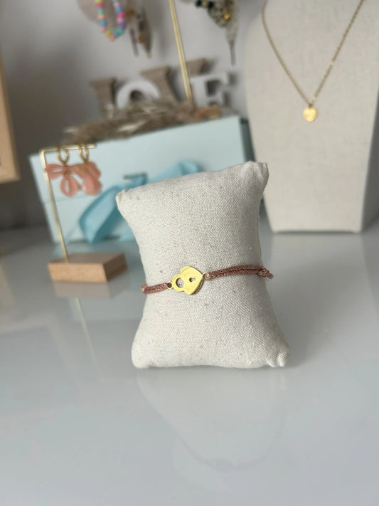 bracelet cordon scintillant saumon à fil doré présenté sur un mini-coussin beige en coton.  Bijou orné d'un cadenas coeur en acier inoxydable doré. En fond un buste avec un collier doré et une paire de boucles d'oreilles dorées et saumon sur un présentoir en bambou. 