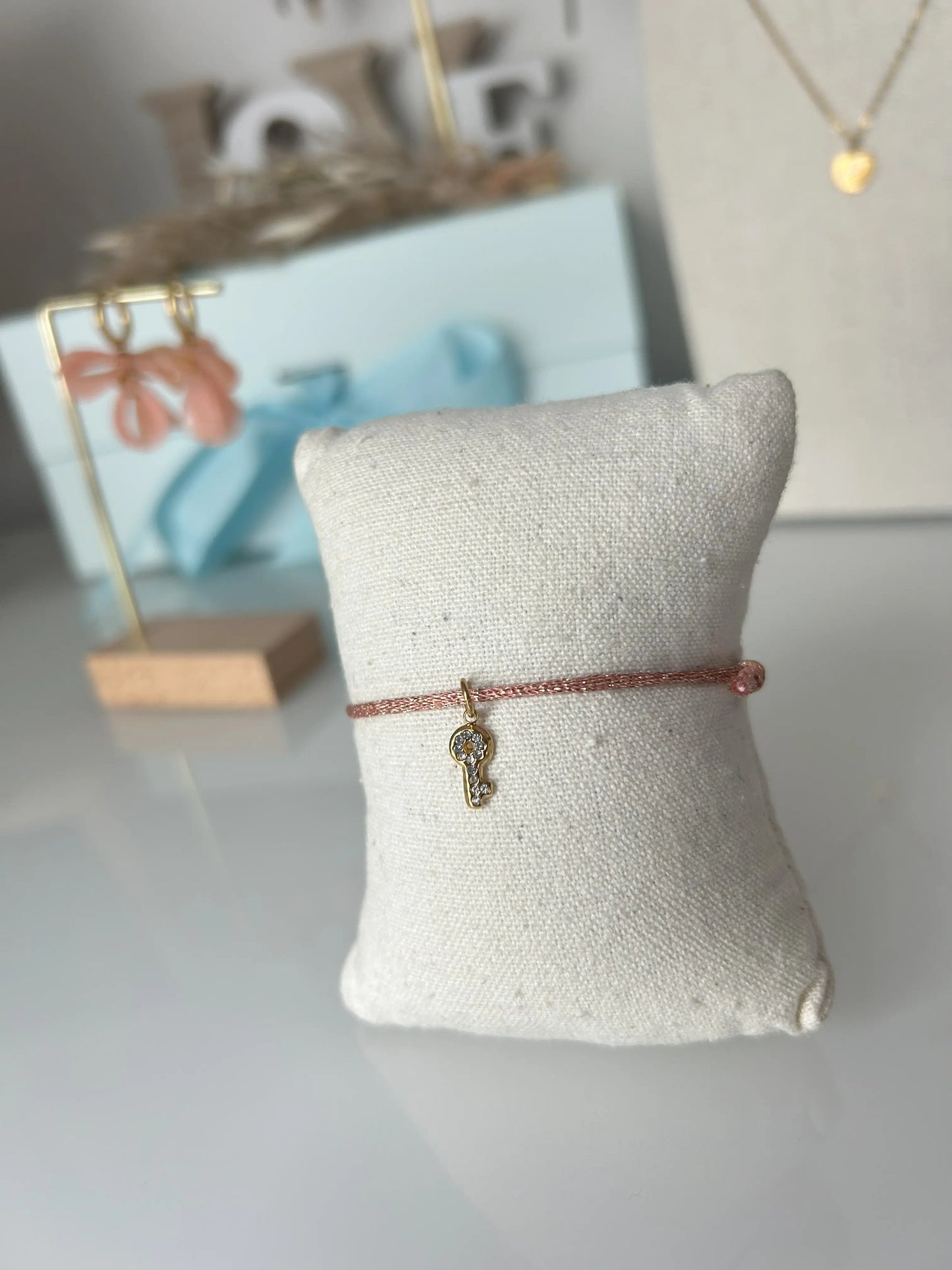 bracelet cordon scintillant saumon à fil doré présenté sur un mini-coussin beige en coton.  Bijou orné d'une clé avec strass en acier inoxydable doré. En fond un buste avec un collier doré et une paire de boucles d'oreilles dorées et saumon sur un présentoir en bambou. 
